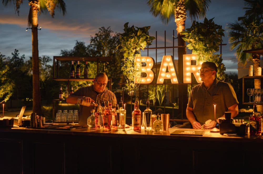 Two bartenders behind the lit BAR sign after dark